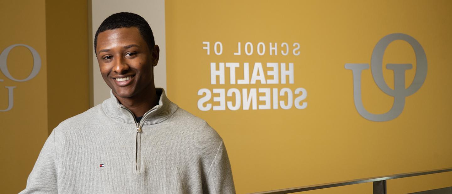 image of a student standing in the Human Health Building in front of a sign with the interlocking OU and School of Health Sciences text